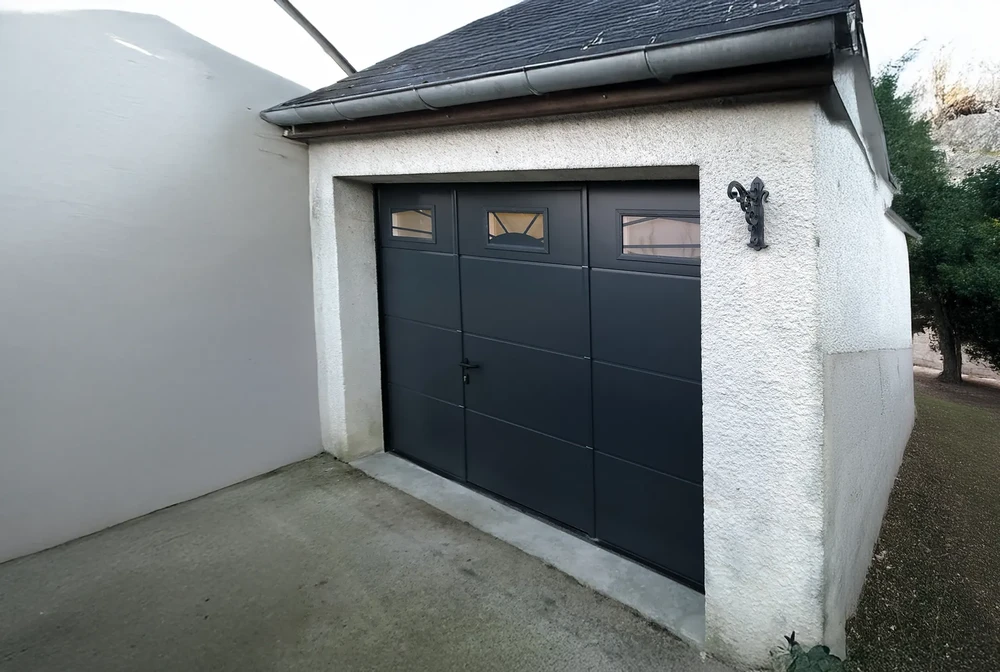 Extérieur résidentiel avec porte de garage sectionnelle anthracite à trois vantaux, équipée de hublots rectangulaires sur les panneaux supérieurs